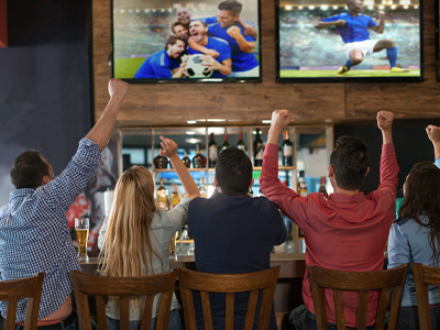 View of a bar with people sitting, their backs facing the camera, watching a sports event on TV