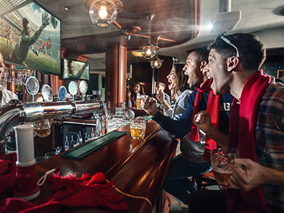 Side view of a bar with people sitting together in sports gear cheering at a TV showing a sports event