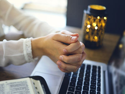A pair of hands clasped in prayer by a laptop and an open bible