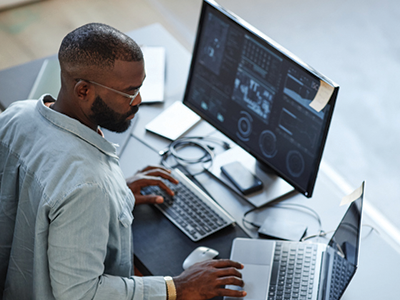 A man sitting at a work desk using a computer and a laptop 