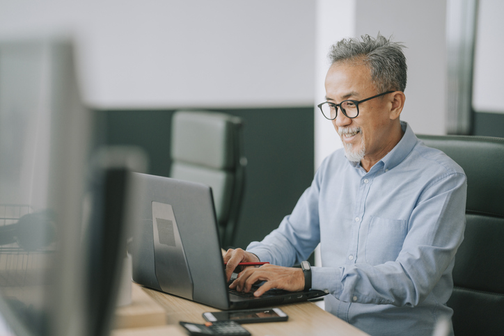 Older Asian Man sitting at a work desk with a laptop