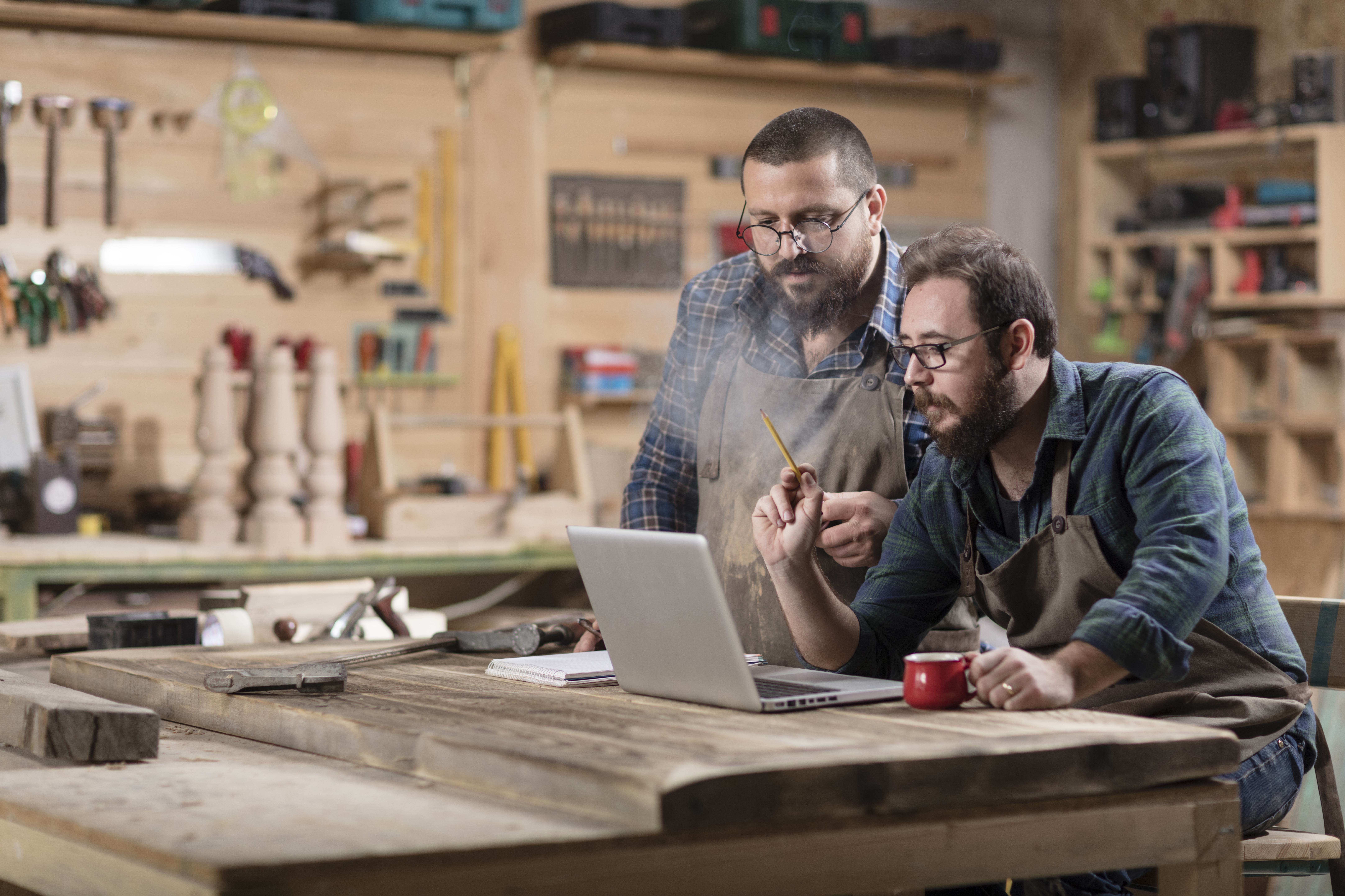 Two white men sitting at a wood/tool workshop on a laptop