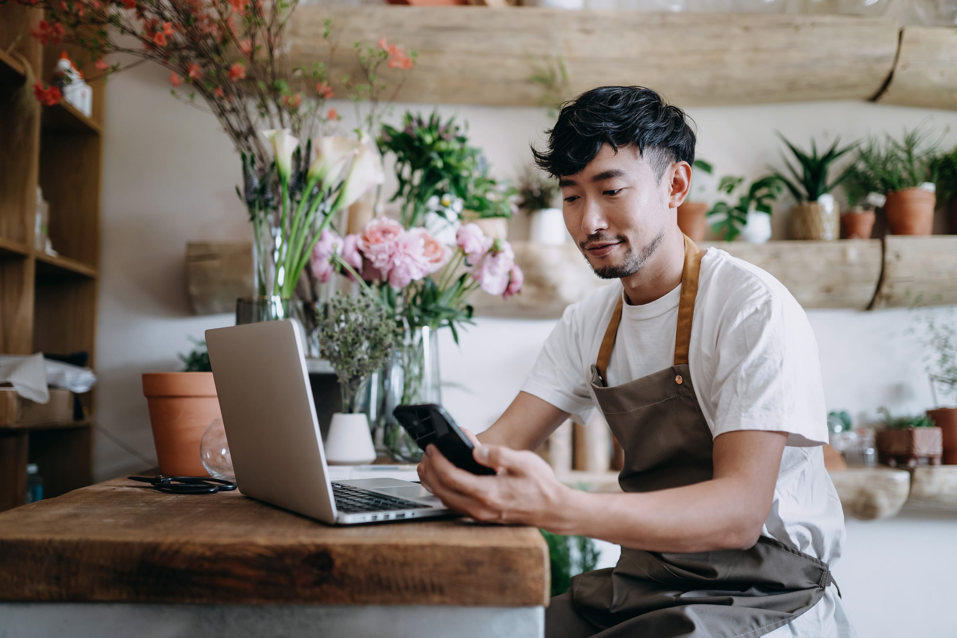 A florist wearing an apron sits at a wooden counter using a smartphone and laptop. Surrounding him are lush plants and flowers, conveying a calm, focused atmosphere.