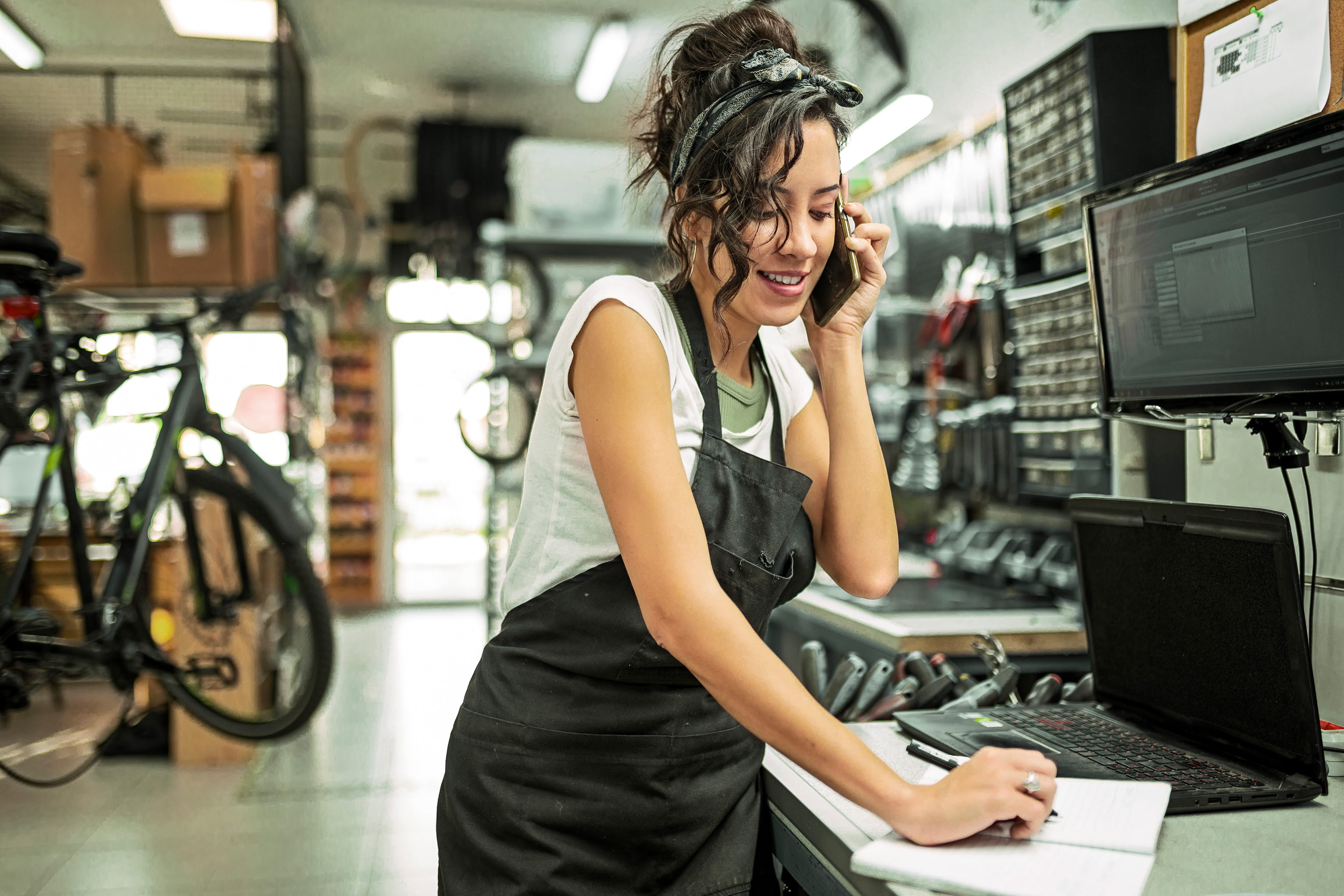 A woman in a bike shop wearing an apron talks on the phone, smiling as she writes in a notebook. Bicycles and repair equipment surround her, creating a busy, lively workspace.
