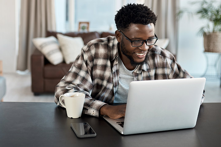African American male sitting in a living room looking at a laptop with a white mug and cellphone next to computer
